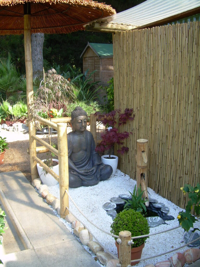 Jardín zen con piedra blanca, estatua de buda, fuente de bambú y vallado de caña natural